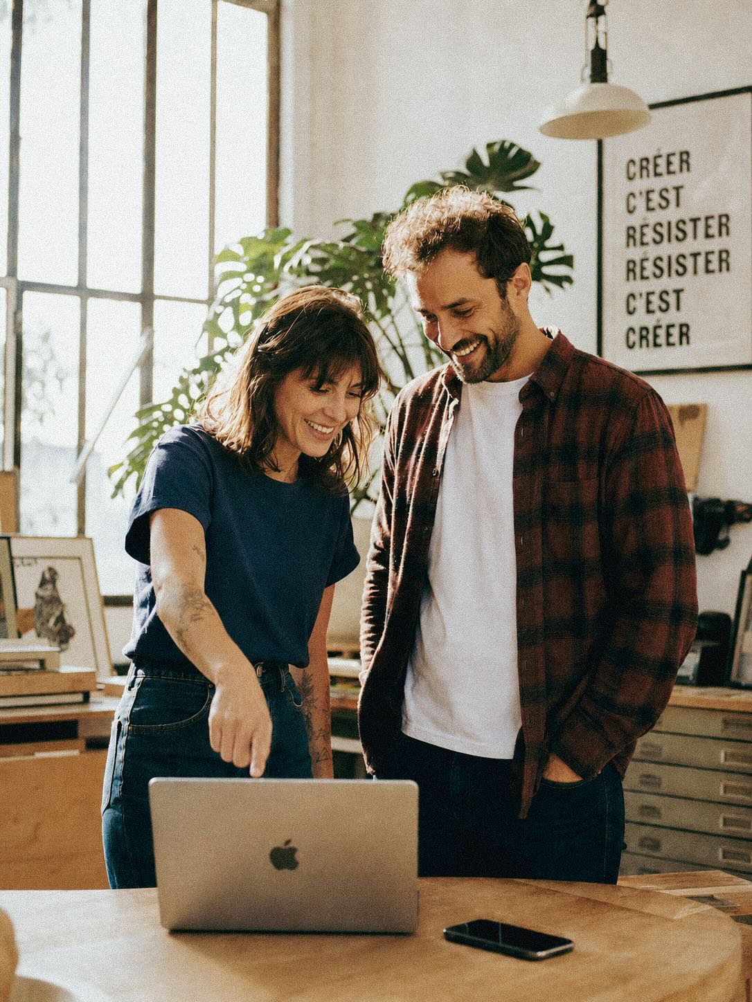 Jeune couple d'entrepreneurs complices devant leur MacBook dans un atelier-loft lumineux à Nantes